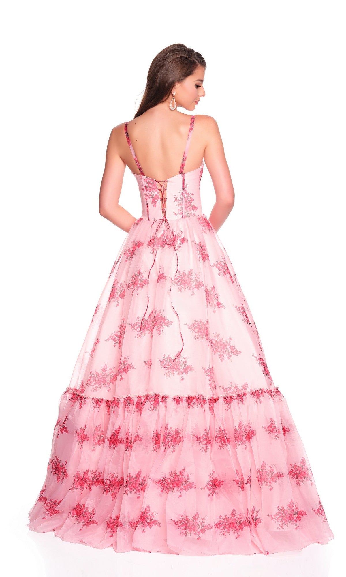 A woman stands facing away in the Long Prom Dress: Dave and Johnny 12934, a pink gown with a satin corset bodice, lace-up back, thin straps, and floral patterns, set against a white background.